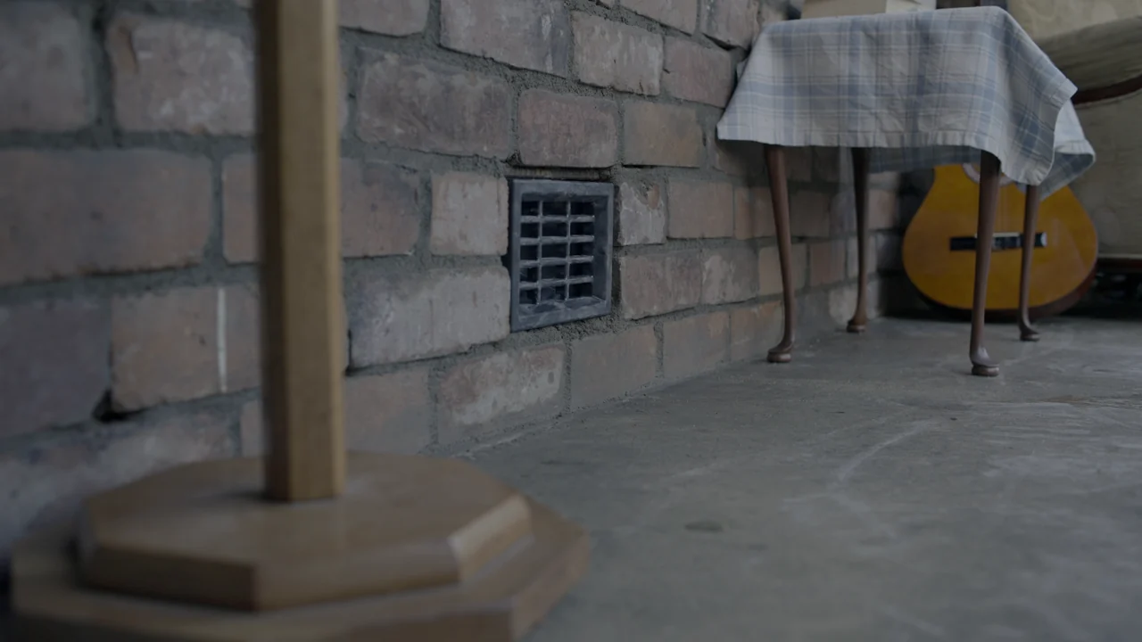 A brick wall with a cast-iron vent, a cloth-covered table and a guitar waiting on the set before the fire